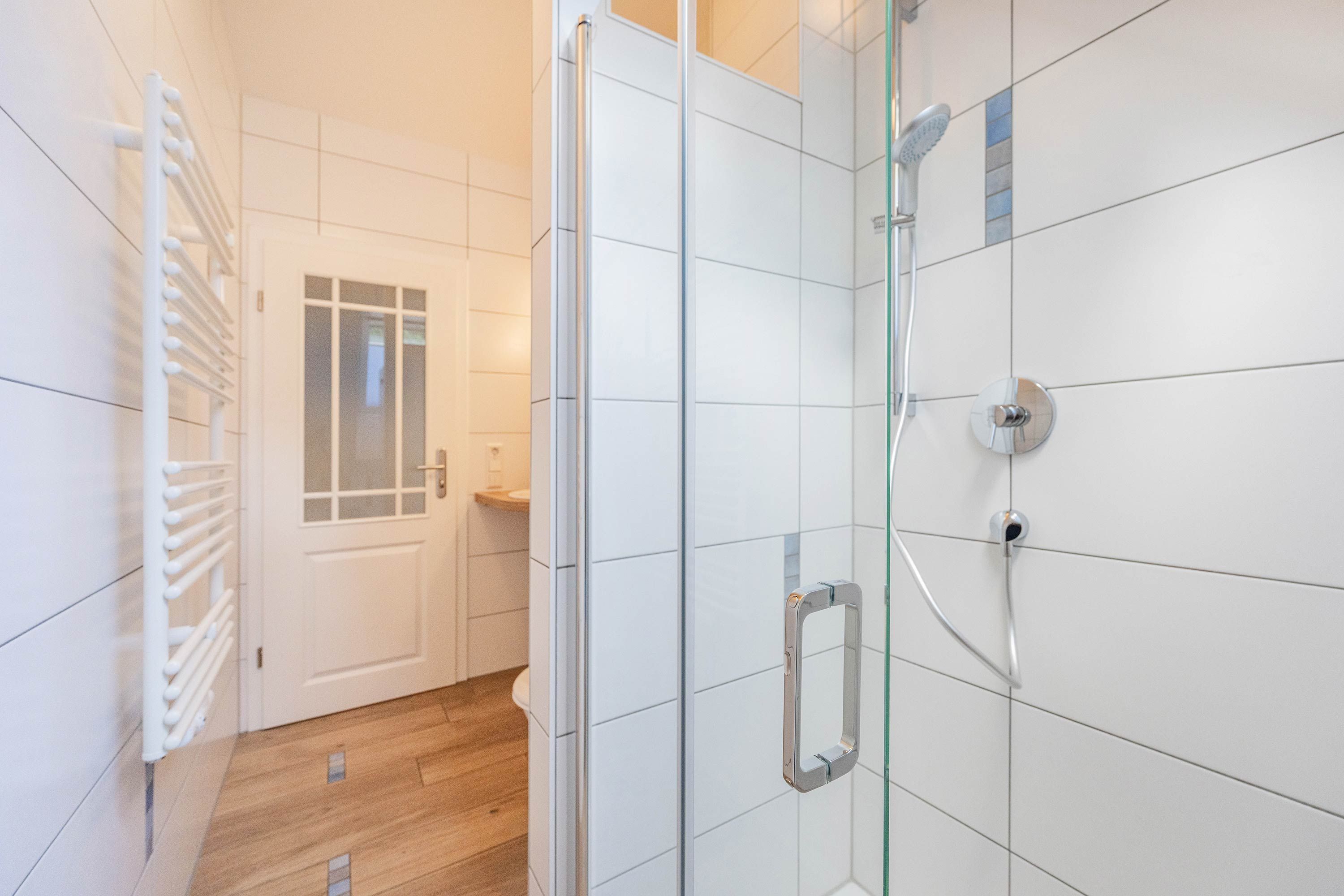 A modern bathroom with white tiles, a glass shower enclosure, and a wooden floor.