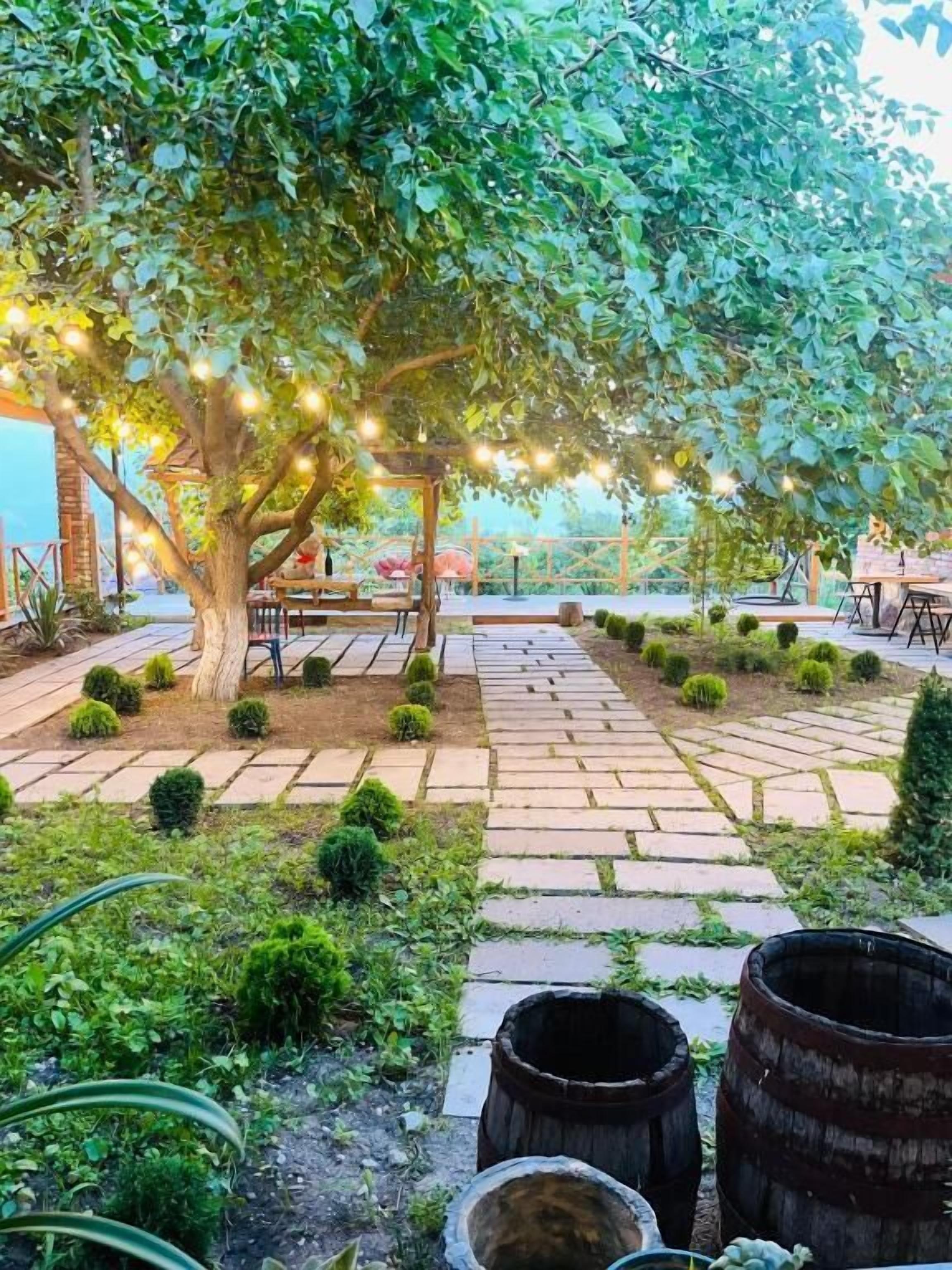 Un jardín exterior con un sendero empedrado, una pérgola de madera con luces colgantes y una zona de estar bajo un árbol.