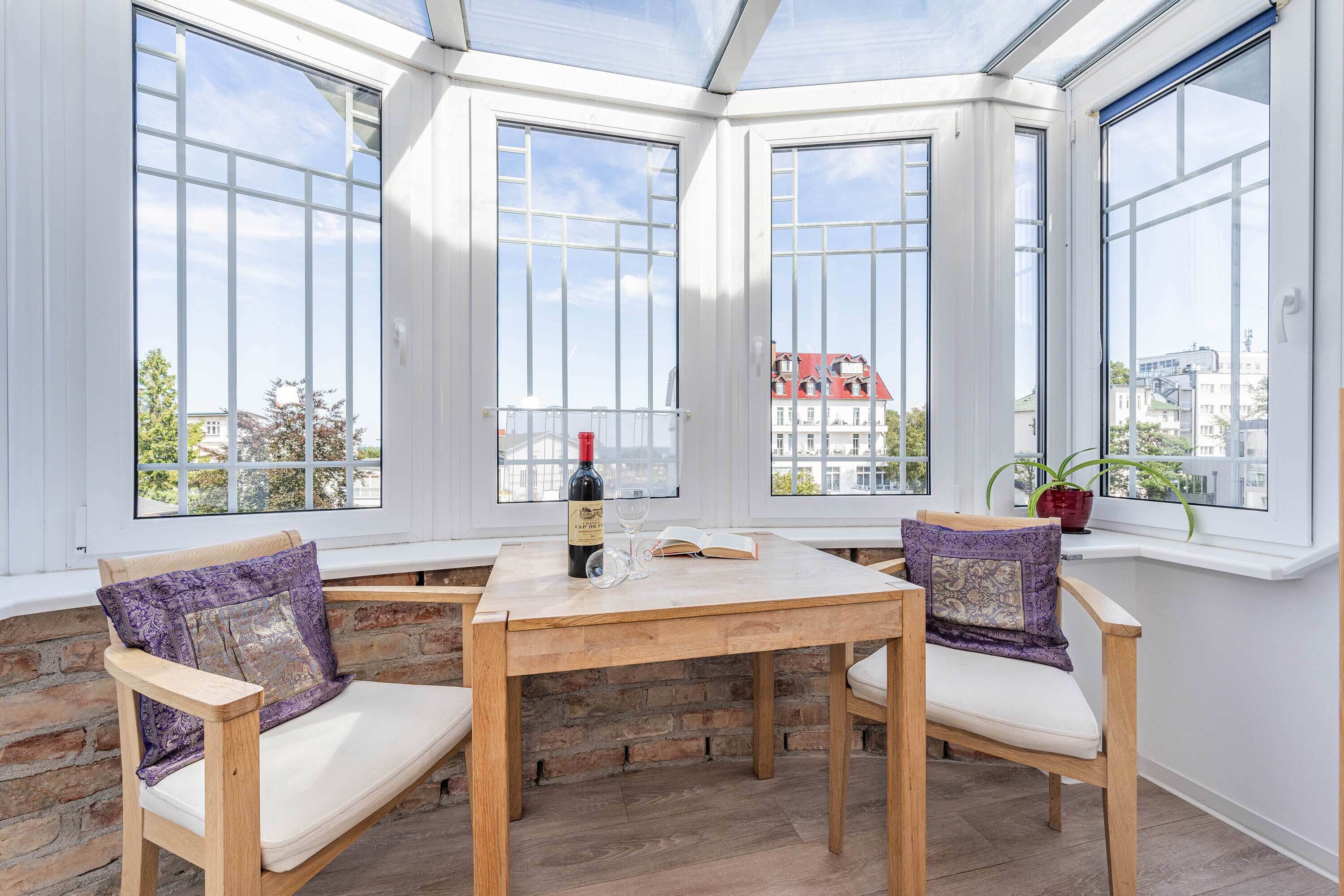 A corner room with large windows, a wooden table, two chairs, and a bottle of wine.