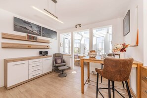 Eine moderne Küche mit weißen Schränken, ein Essbereich mit Holztisch und -stühlen sowie große Fenster für natürliches Licht.