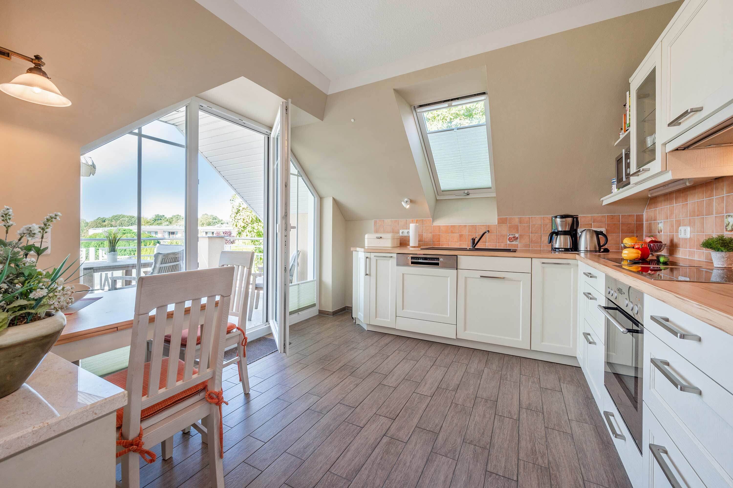 Une cuisine moderne avec des meubles blancs, un coin repas avec une table et des chaises en bois, et une grande porte-fenêtre coulissante donnant sur une terrasse extérieure.