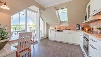 Une cuisine moderne avec des meubles blancs, un coin repas avec une table et des chaises en bois, et une grande porte-fenêtre coulissante donnant sur une terrasse extérieure.