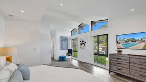 Quarto de hotel moderno com uma cama grande, penteadeira de madeira, um banquinho azul e vista para uma piscina através de grandes janelas.