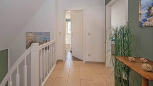 Un pasillo con barandilla blanca, una mesa de madera con adornos, una pared verde con un cuadro y una puerta que da a otra habitación.