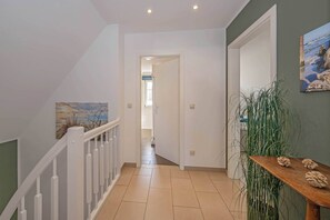 A hallway with a white railing, a wooden table with decorative items, a green wall with a painting, and a door leading to another room. - Modern vacation home with a sauna, garden, and beach access on Usedom (Koserow)