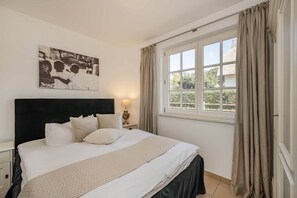 Ein Schlafzimmer mit einem Bett, einem Nachttisch, einer Lampe, einem Fenster mit Vorhängen und einem Wandbild.