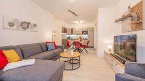 A modern living room with a grey sofa, a dining area with red chairs, a kitchen in the background, and a TV on the right. - Modern vacation rental in Kölpinsee with a balcony—just 600 m from the beach (Ostseebad Kölpinsee)