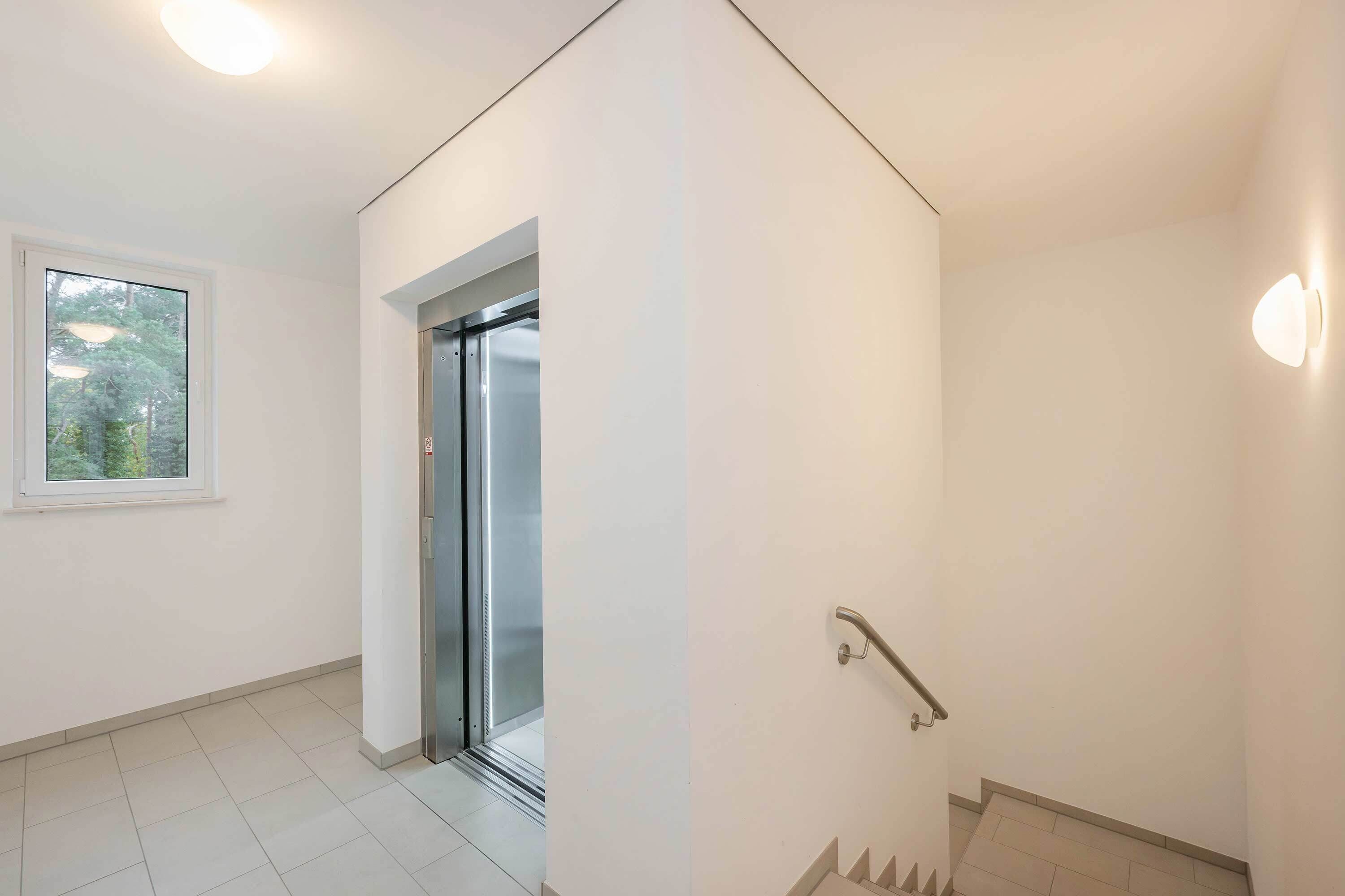 Un interior moderno con una escalera, una puerta que da a un balcón y un ventanal con vistas a árboles.