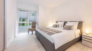 A bedroom with a bed, bedside table, chair, and a window with blinds. - Modern vacation rental in Kölpinsee with a balcony—just 600 m from the beach (Ostseebad Kölpinsee)