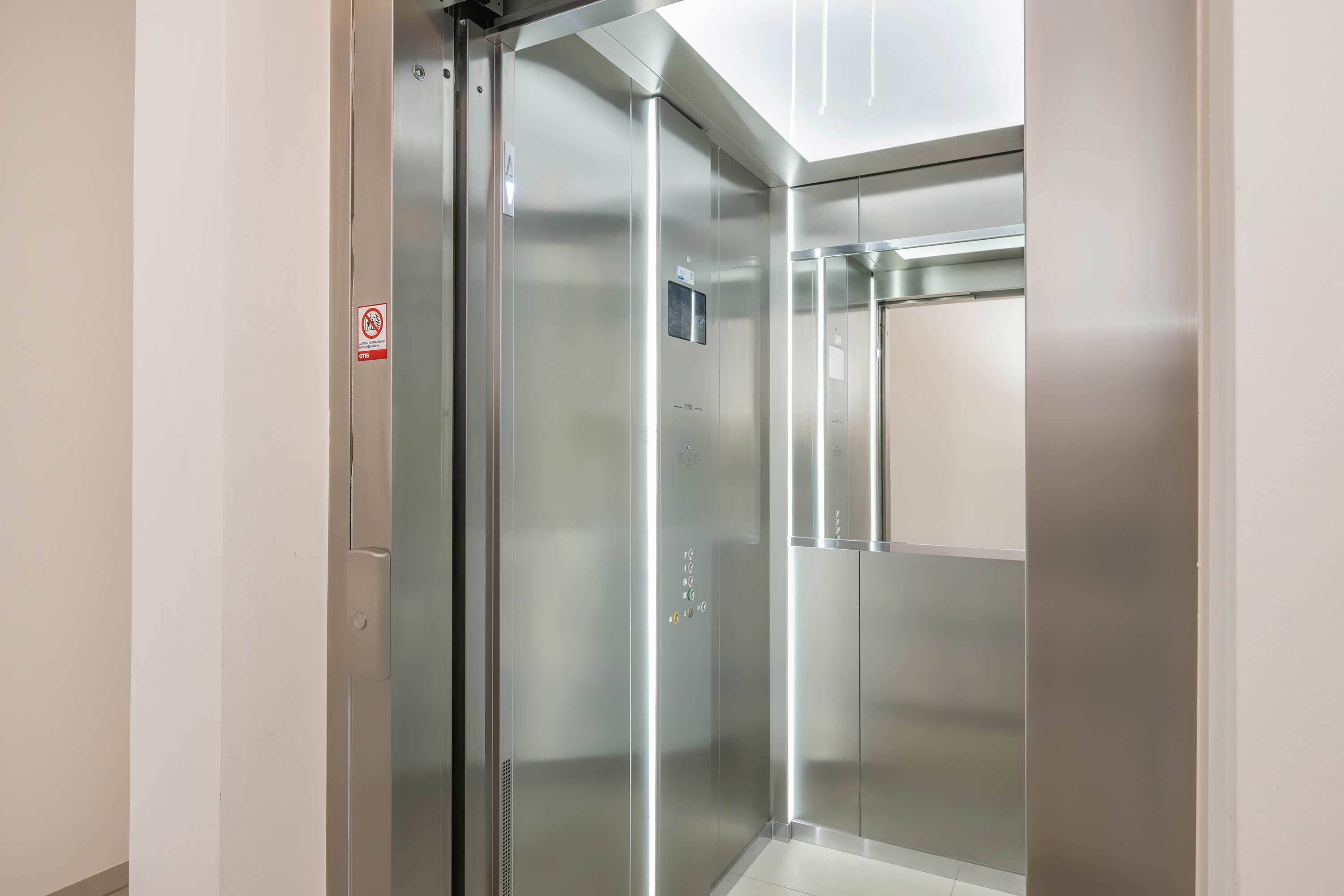 En moderne elevator med metalvægge og en glasdør, med et skilt, der angiver røgfri zone, samt et kontrolpanel med knapper.