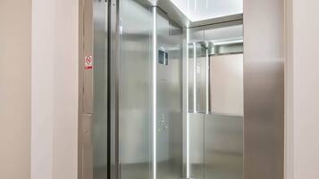 En moderne elevator med metalvægge og en glasdør, med et skilt, der angiver røgfri zone, samt et kontrolpanel med knapper.