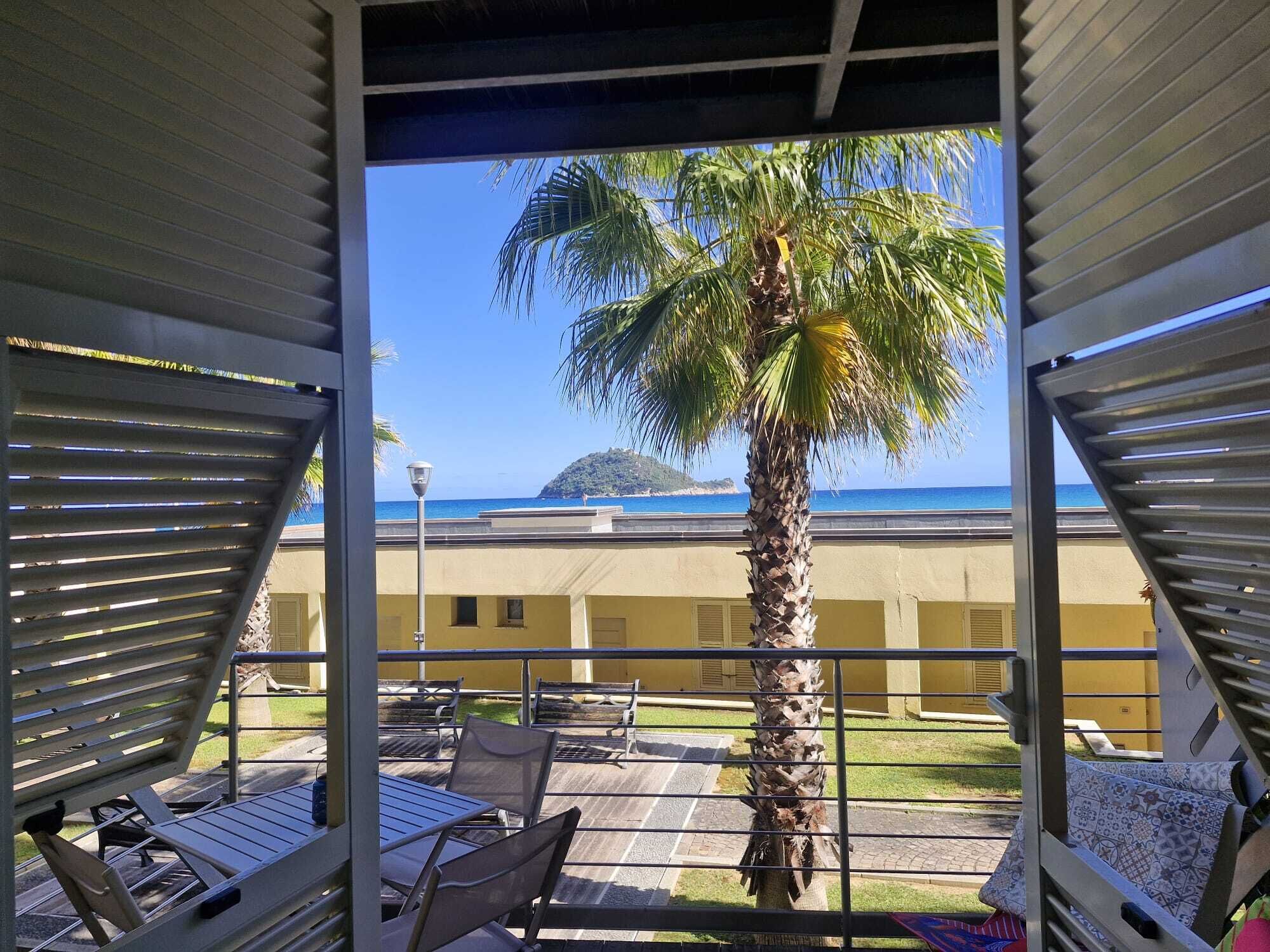 A balcony with a view of the sea, palm trees, and a building in the background.
