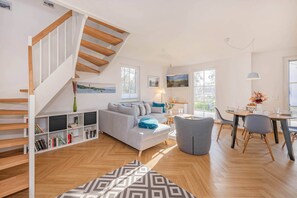 A modern living room with a staircase, a sofa, a dining table, and a bookshelf. - Modern vacation home with a sauna and garden—quiet and comfortable in a green setting (Zirchow)