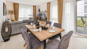 A modern living room with a sofa, a dining table set with plates and glasses, and a large window with curtains. - Modern apartment with a balcony—close to the beach and family-friendly (Ostseebad Kölpinsee)