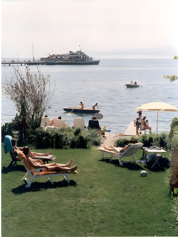 A grassy area with lounge chairs and umbrellas, a dock with boats, and a large ship in the background. - Studio 'Typ A 18' with Lake View, Shared Garden and Wi-Fi (Immenstaad)