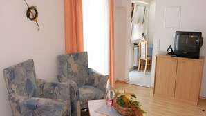 Una sala con dos sillones estampados, una mesita con una cesta decorativa, un televisor sobre un mueble de madera y un acceso a otra habitación.