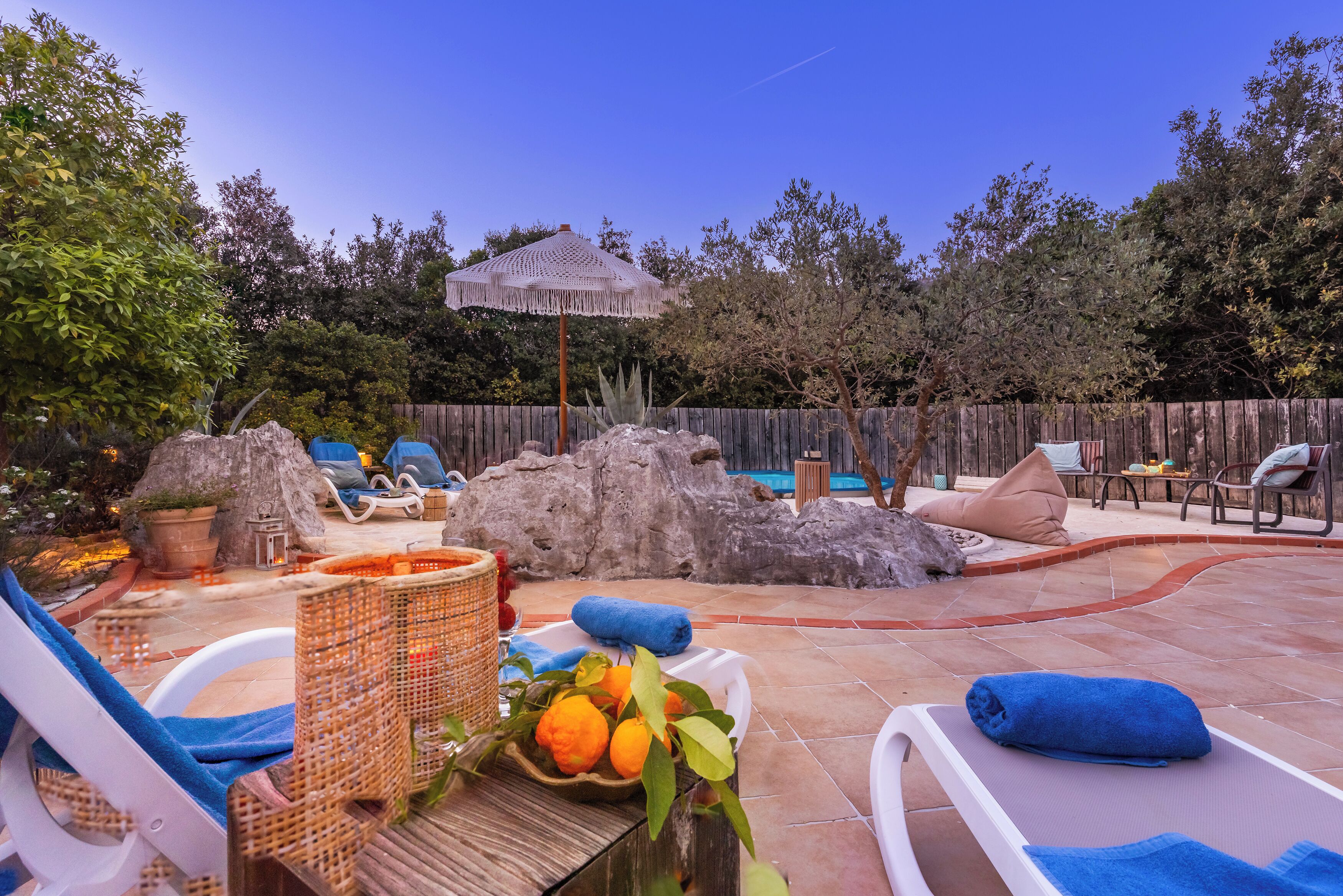 Un jardin avec une piscine, des chaises longues, un parasol de style tiki et un élément décoratif en pierre.