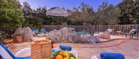 Ein Garten mit Pool, Liegestühlen, einem Tiki-Sonnenschirm und einem Steinelement.