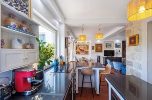A modern kitchen with a coffee machine, bar stools, and a dining area with a stone wall and artwork. - Charming 4-Bedroom Stone Villa with Pool & Garden, 15 Min to Dubrovnik (Knežica)