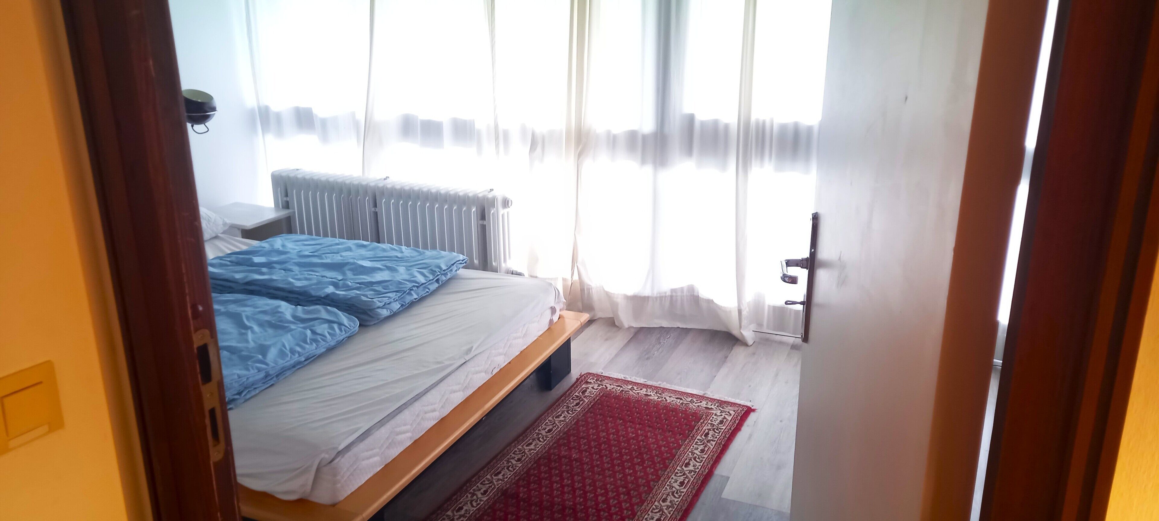 Un dormitorio con cama, radiador, alfombra roja y ventana con cortinas.