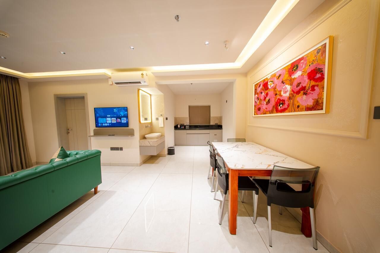 Een moderne woonkamer met een groene bank, een eettafel met stoelen, een televisie, een keukenhoek en een groot bloemen schilderij aan de muur.