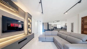 En moderne stue med et stort fladskærms-tv, en hjørnesofa og et køkken i baggrunden.