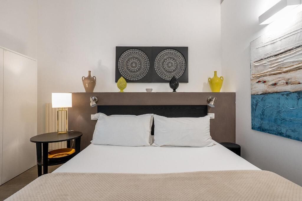 A hotel room with a bed, bedside tables, a lamp, decorative vases, and a wall painting.