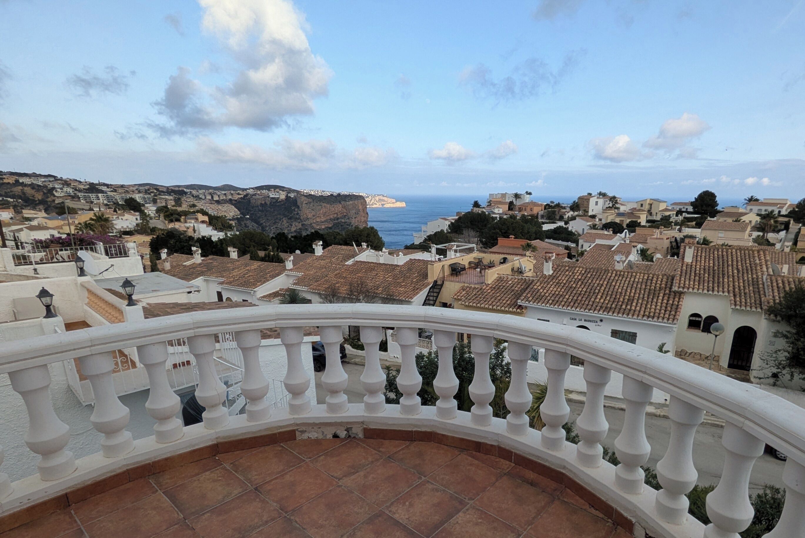 Un balcon doté d’une balustrade blanche, qui offre une vue sur une ville côtière aux toits de tuiles rouges, sous un ciel dégagé.