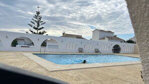 Une piscine où une personne nage, entourée de bâtiments blancs dotés d’arcades, sous un ciel dégagé.