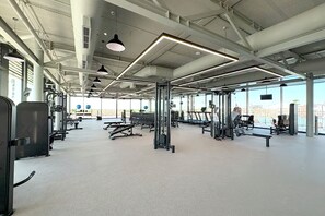 A spacious gym with various exercise equipment, large windows, and a high ceiling.