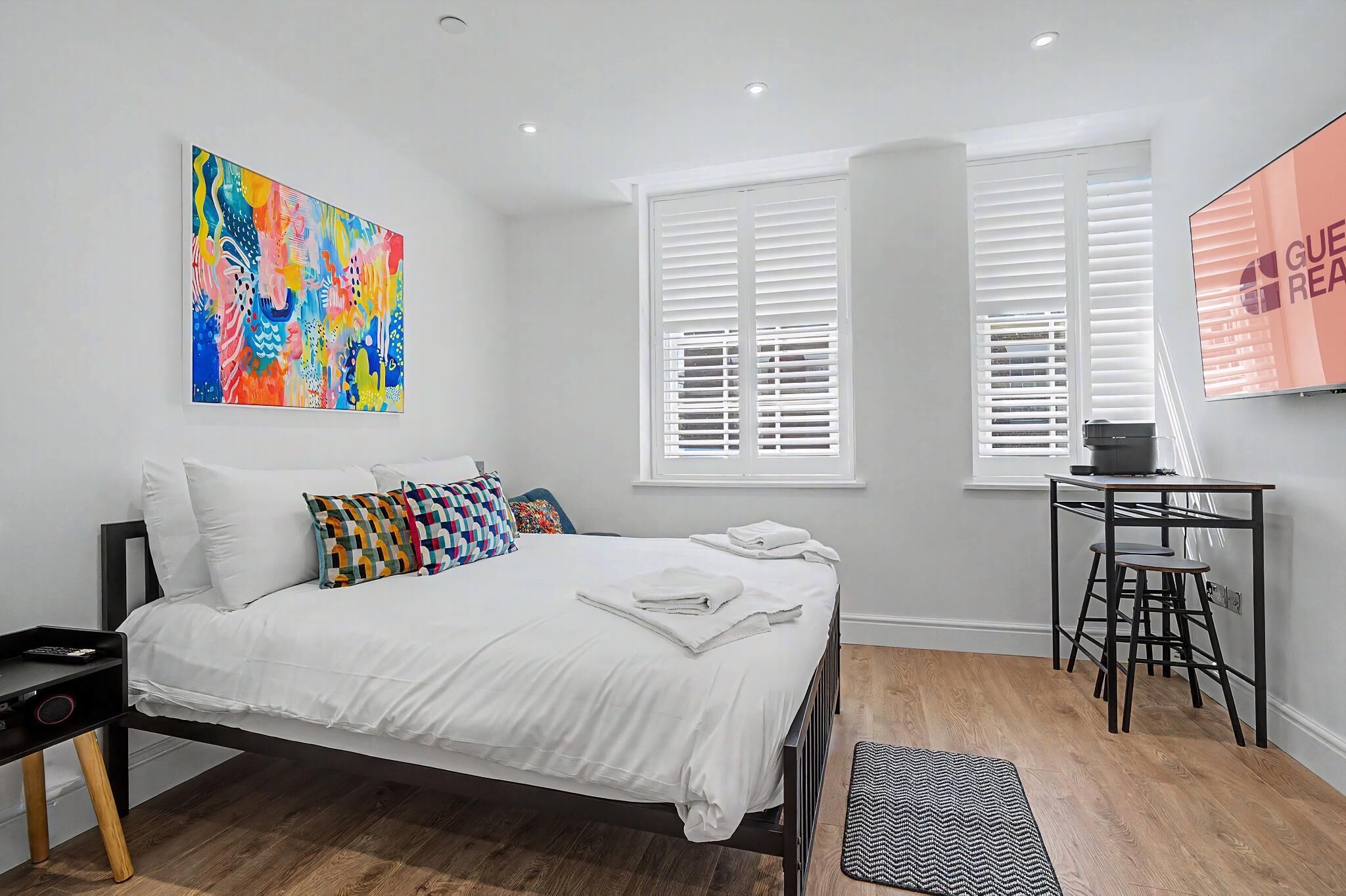 A modern bedroom with a bed, a colorful abstract painting, a desk with a chair, and a window with blinds.