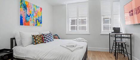 A modern bedroom with a bed, a colorful abstract painting, a desk with a chair, and a window with blinds.