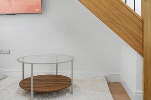 A small round table with a glass top and wooden base, positioned on a white rug in a room with white walls and a wooden staircase. - Guestready - Duke Street (Liverpool)