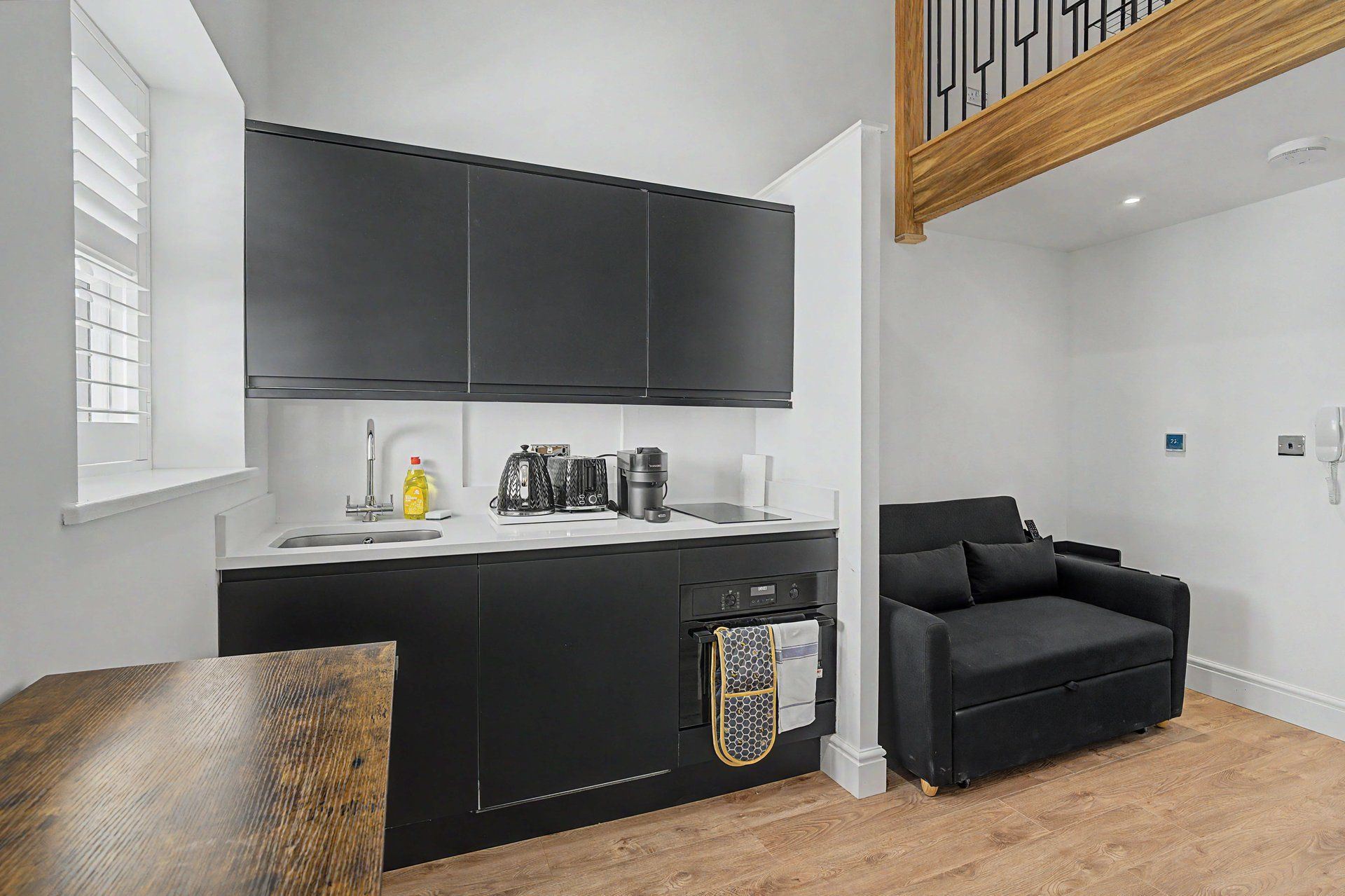Cocina compacta con armarios negros, encimera blanca y sofá negro en un apartamento moderno.