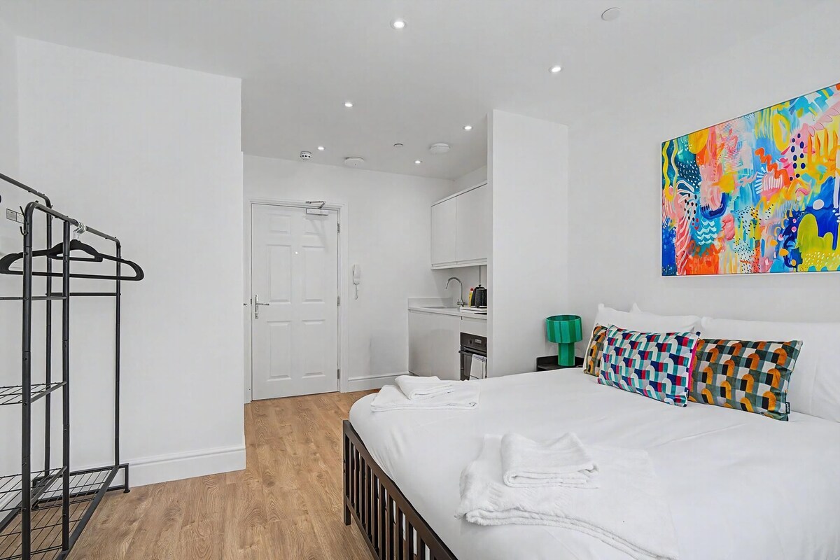 A hotel room with a bed, a wardrobe, a kitchenette, and a colorful abstract painting.