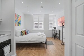 Un dormitorio con cama, escritorio y silla, una ventana con persianas y un cuadro colorido en la pared.