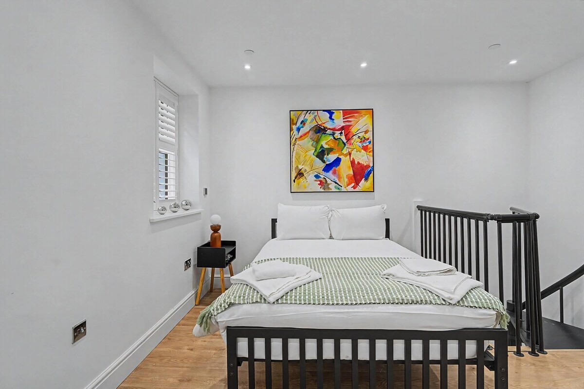 A bedroom with a bed, a window with blinds, a nightstand, a colorful abstract painting, and a black railing.