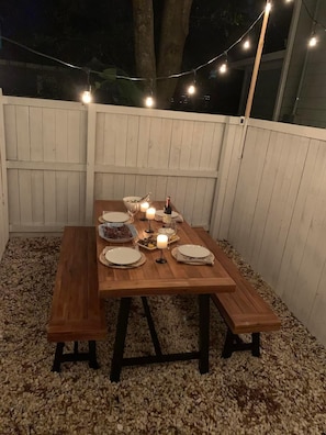Una mesa de madera al aire libre dispuesta para comer, con velas, platos y una botella de vino, rodeada por una cerca blanca y luces colgantes.