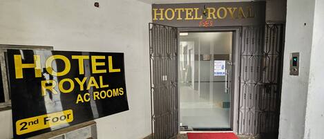 A hotel entrance with a sign for 'HOTEL ROYAL AC ROOMS' on the wall and a red carpet leading to the door.