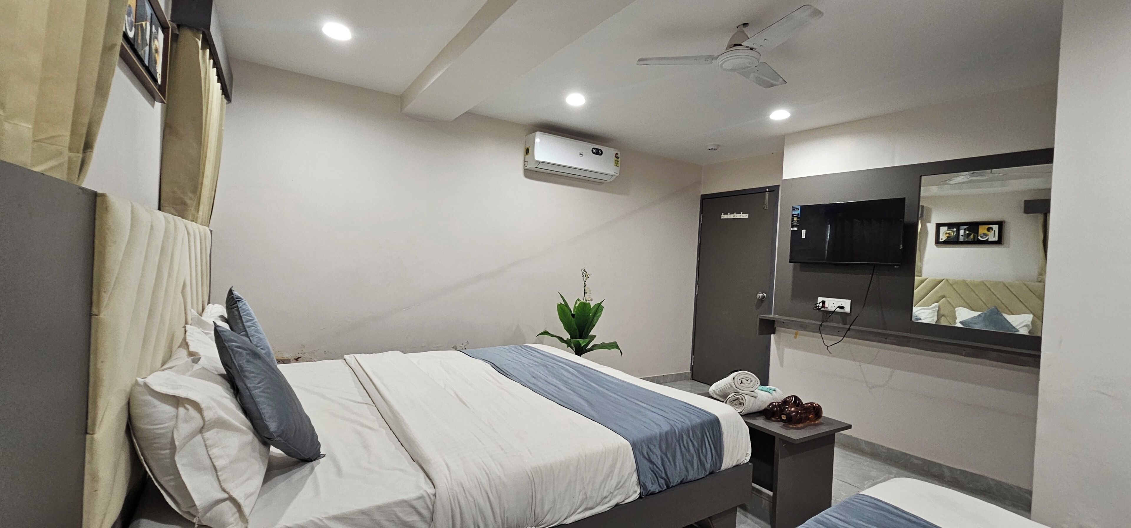 Une chambre d’hôtel équipée d’un lit, d’une table de chevet, d’un climatiseur, d’un ventilateur de plafond, d’une télévision et d’un miroir.