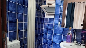 Ein Badezimmer mit blauen Fliesen, einer weißen Toilette, einem Duschvorhang und einem Waschbecken mit Pflegeprodukten.