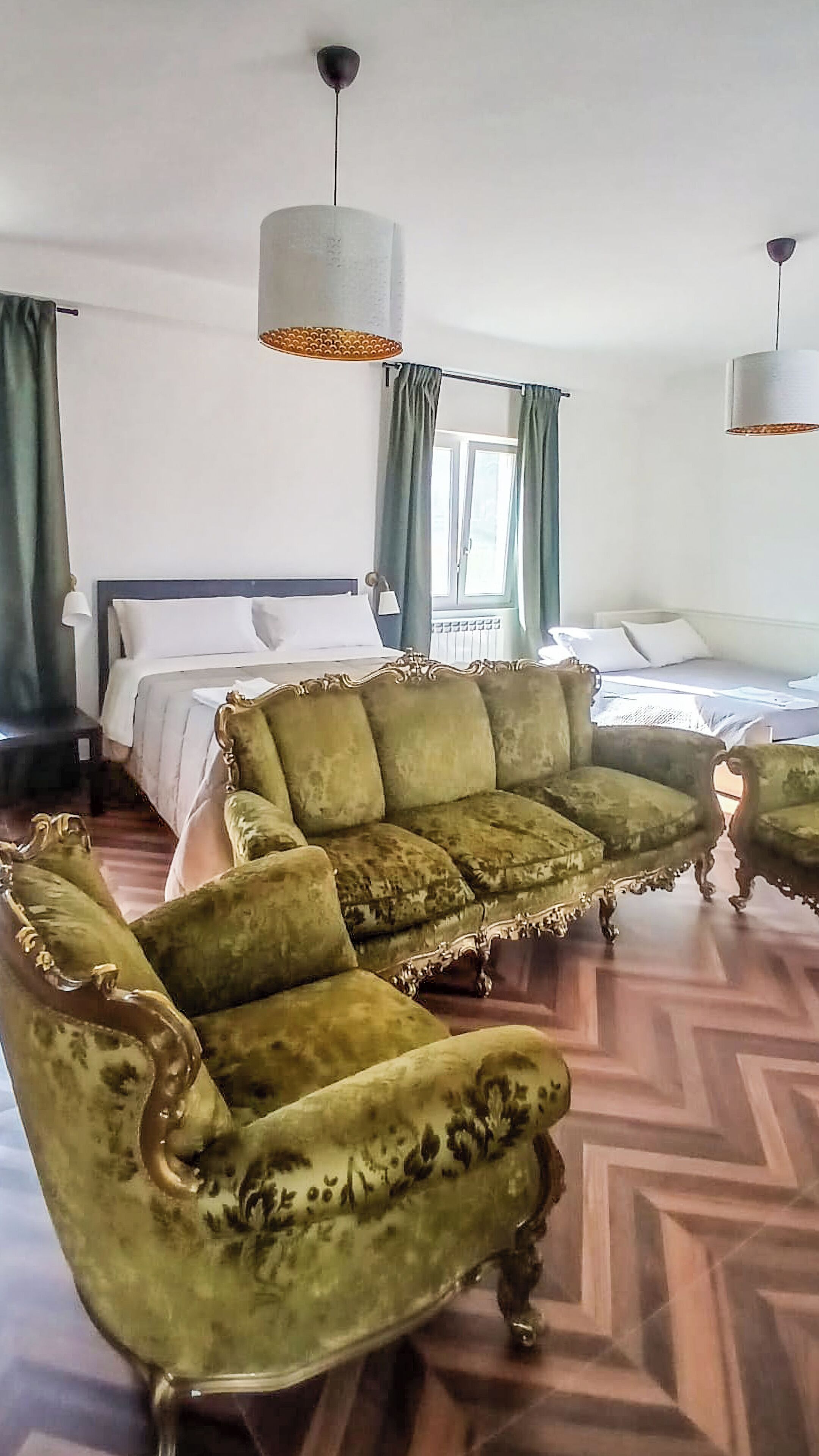 A room with a large green sofa, two matching armchairs, a bed with white linens, and two pendant lights.