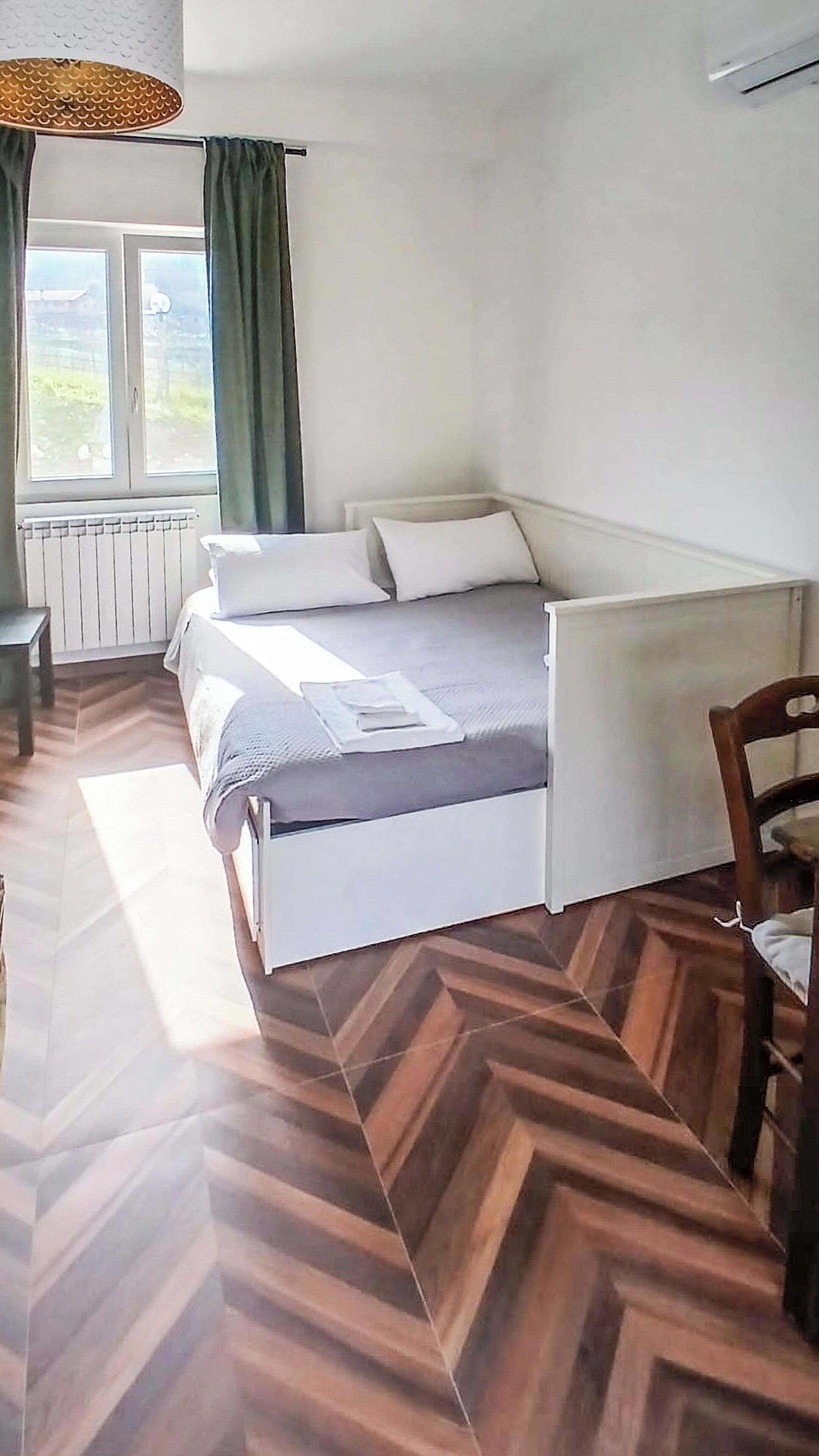 A bedroom with a bed, a chair, a table, a window with curtains, and a radiator.