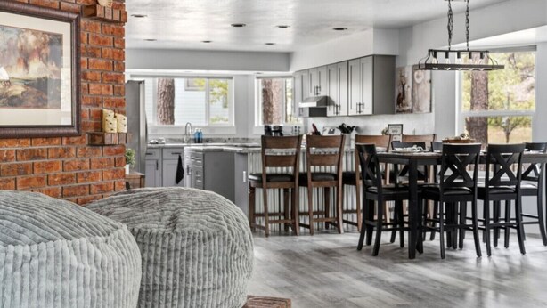 A modern kitchen with a dining area, featuring a brick wall, wooden chairs, and a chandelier.