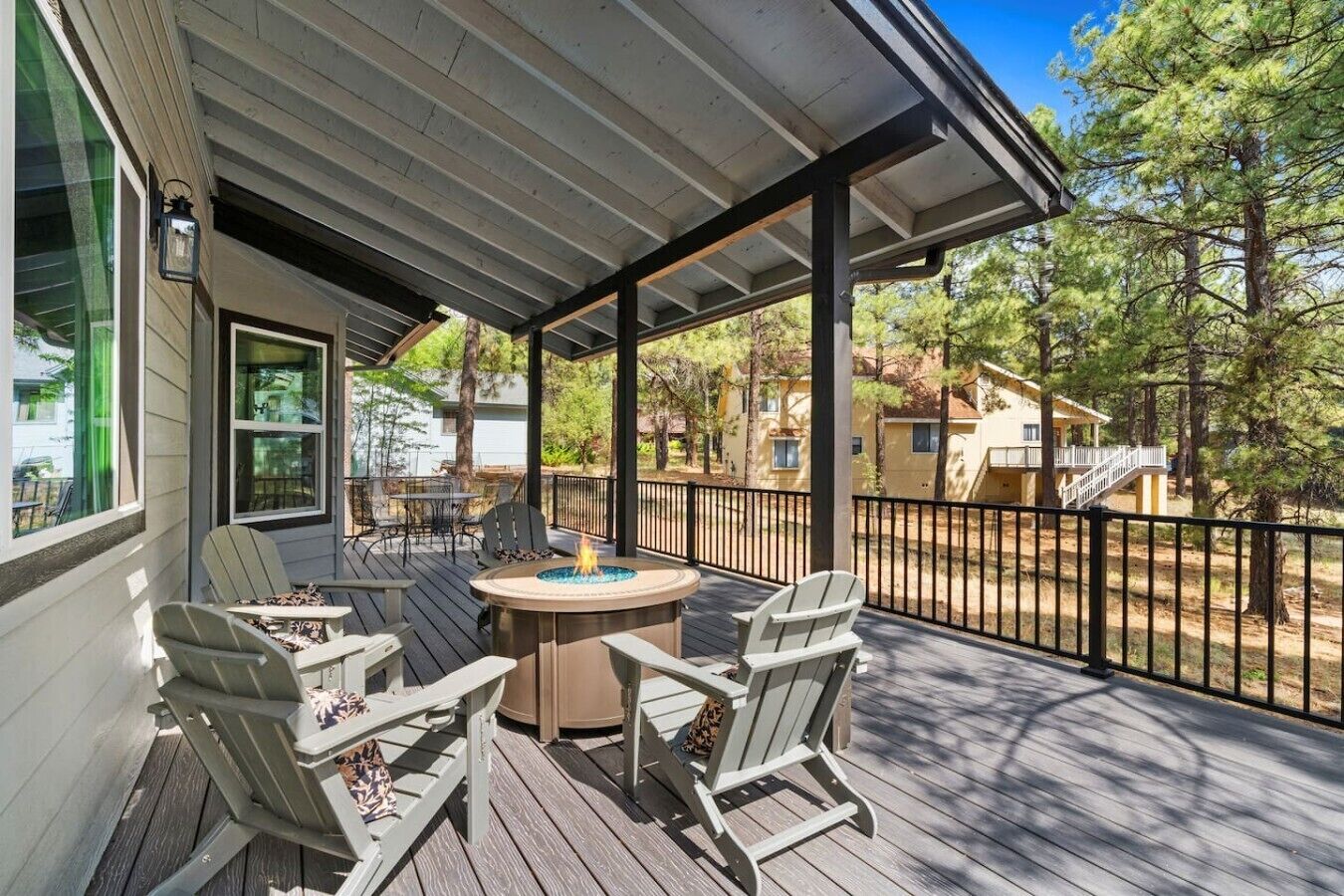A covered deck with outdoor furniture, a fire pit, and a view of surrounding trees and buildings.