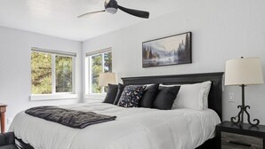 A bedroom with a large bed, a ceiling fan, a nightstand with a lamp, and a painting on the wall.