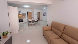 A modern living room with a sofa, a dining area with chairs and a table, and a kitchen in the background.