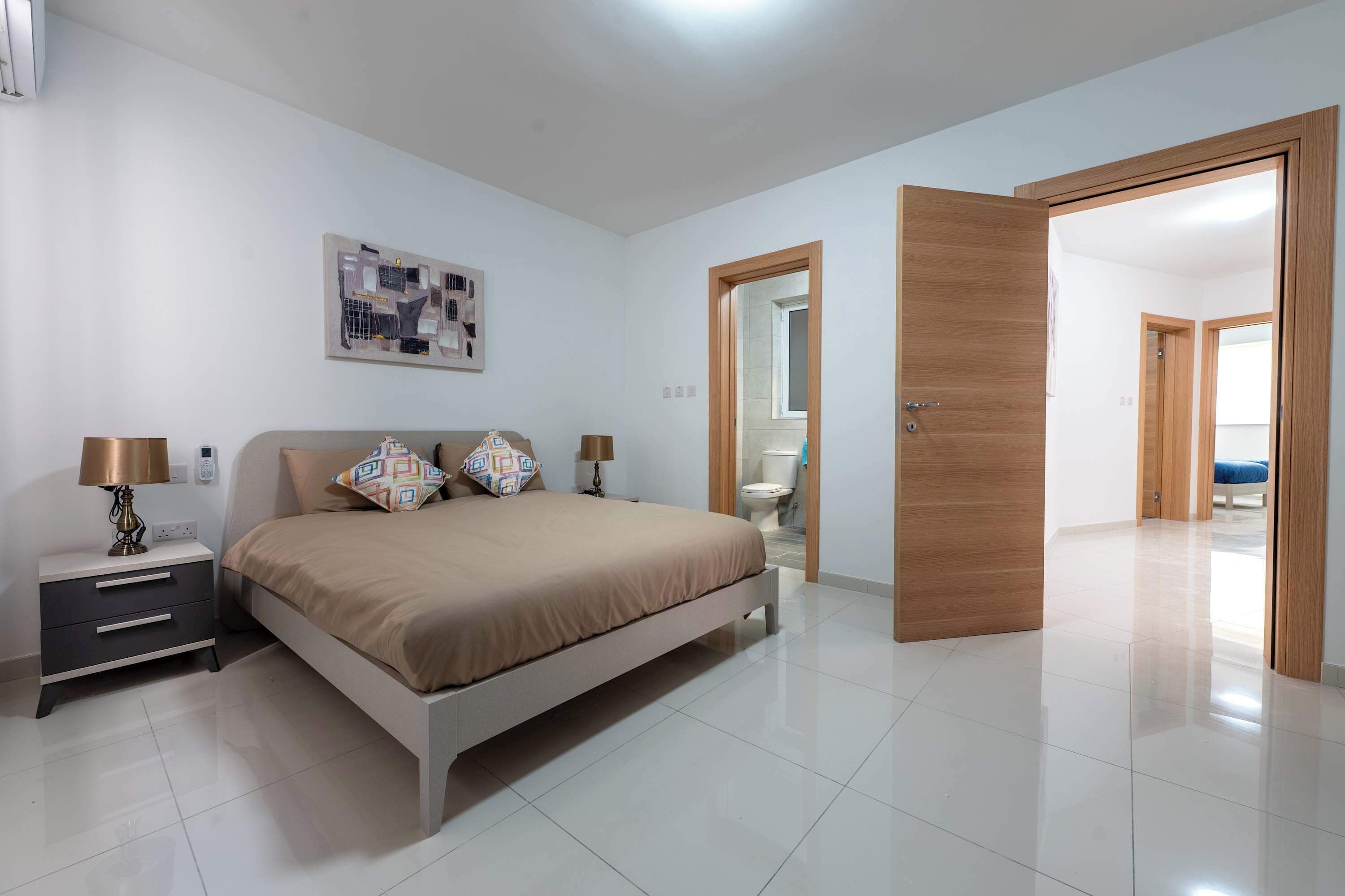 A bedroom with a bed, bedside tables, a wall-mounted artwork, and an open door leading to another room.