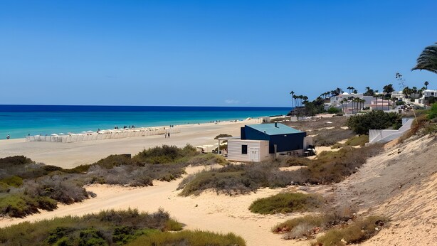 A coastal beach area with a clear sky, sandy terrain, and a small building with a dark roof. - Casa Bella 1 - Apartment with Sea and Beach Views (Costa Calma)