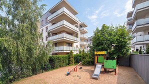 A playground with a seesaw and slide, surrounded by a sandy area and trees, with multi-story residential buildings in the background. - Spacious Apartment with a Balcony | Parking | 2 Bedrooms (Toruń)